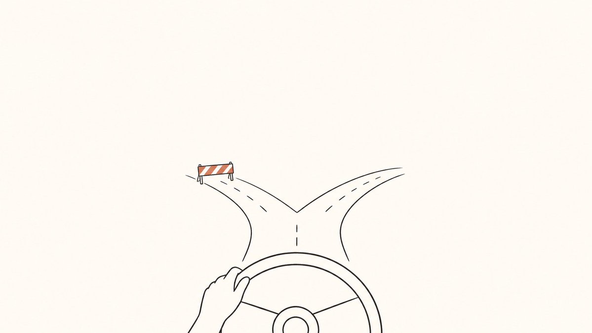 A hand on a steering wheel faces a fork in the road, with a barrier blocking one branch.