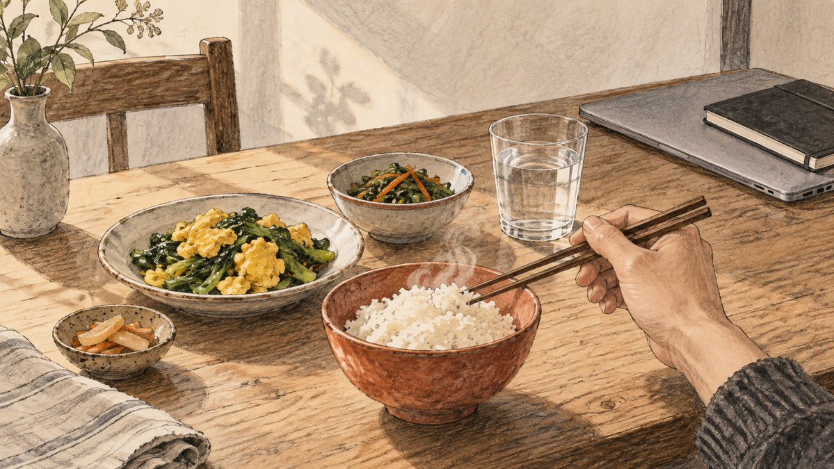 A hand holds chopsticks over a meal beside a glass of water, a closed laptop, and a notebook.